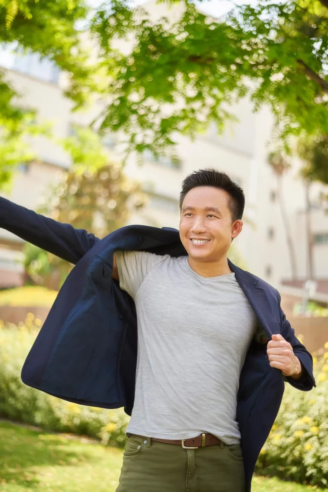 Bright outdoor dating photo for men in Los Angeles, smiling man in a gray t-shirt putting on a navy jacket in a green outdoor setting, natural lifestyle image for an online dating profile.