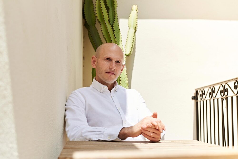 Man seated outdoors in a white button-down shirt, relaxed dating profile portrait with direct eye contact, cactus plants, and bright architectural background