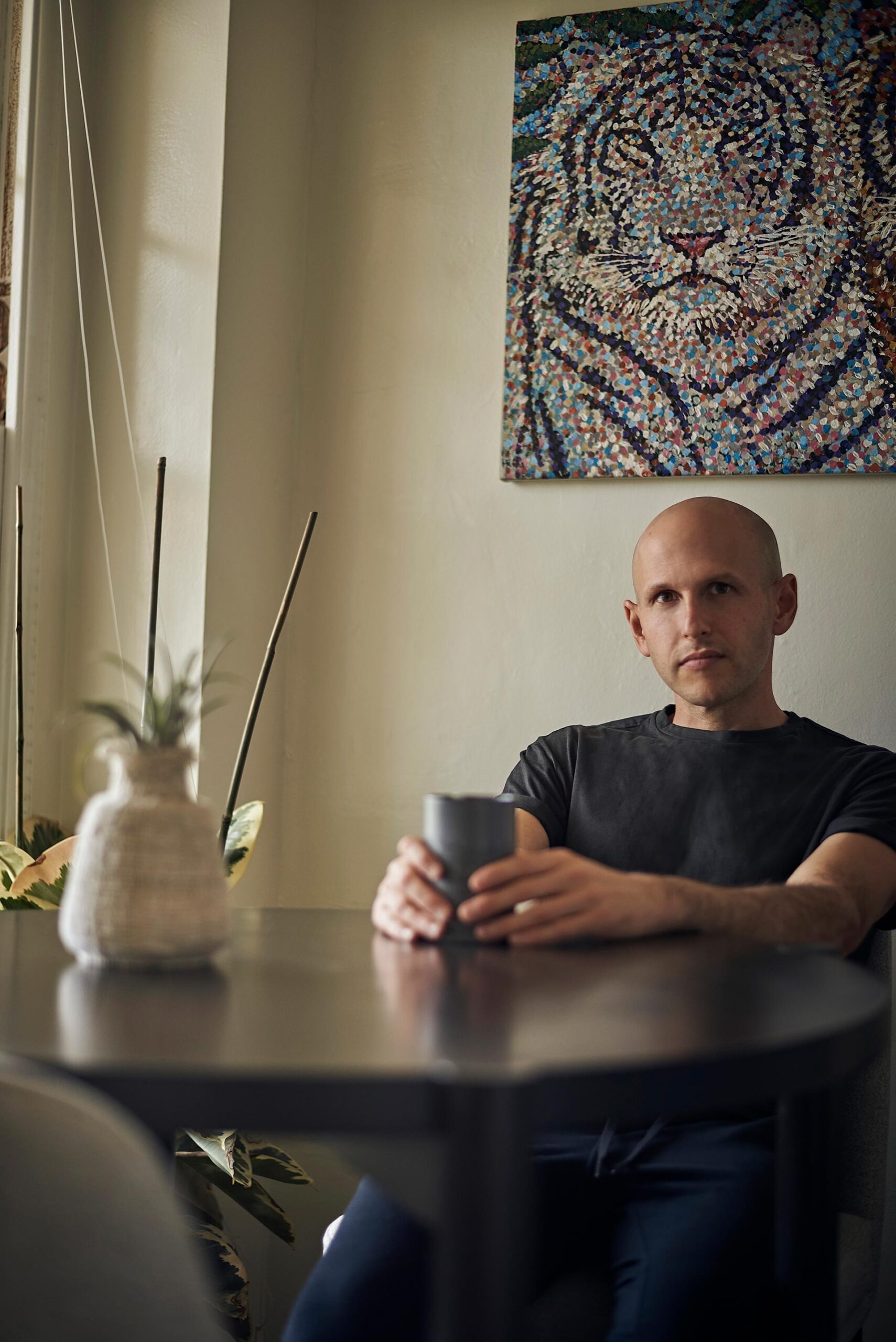 Man seated indoors at a cafe table holding a mug, casual dating profile portrait with soft window light, direct eye contact, dark shirt, and artwork in the background