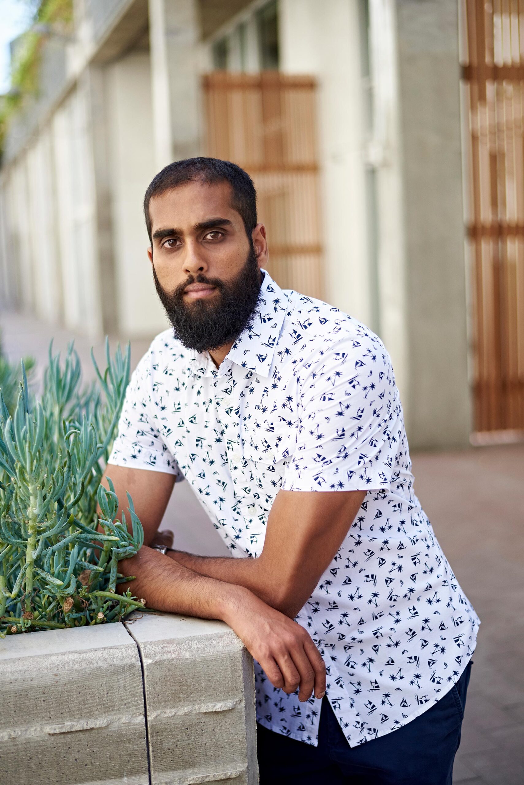 Man leaning against a planter outdoors, casual dating profile portrait with patterned short-sleeve shirt, direct eye contact, greenery, and modern walkway background