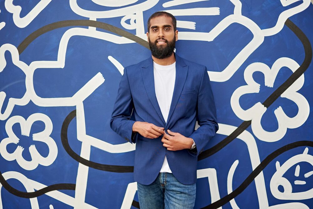 Man standing in front of a blue mural, dating profile portrait with blue blazer, white shirt, jeans, direct eye contact, and a confident relaxed pose