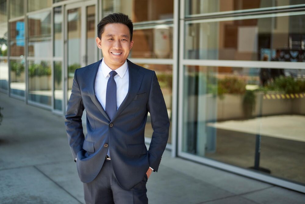 Man in a navy suit and tie standing outside a modern glass building, polished dating profile portrait with natural smile, hands in pockets, and clean architectural background