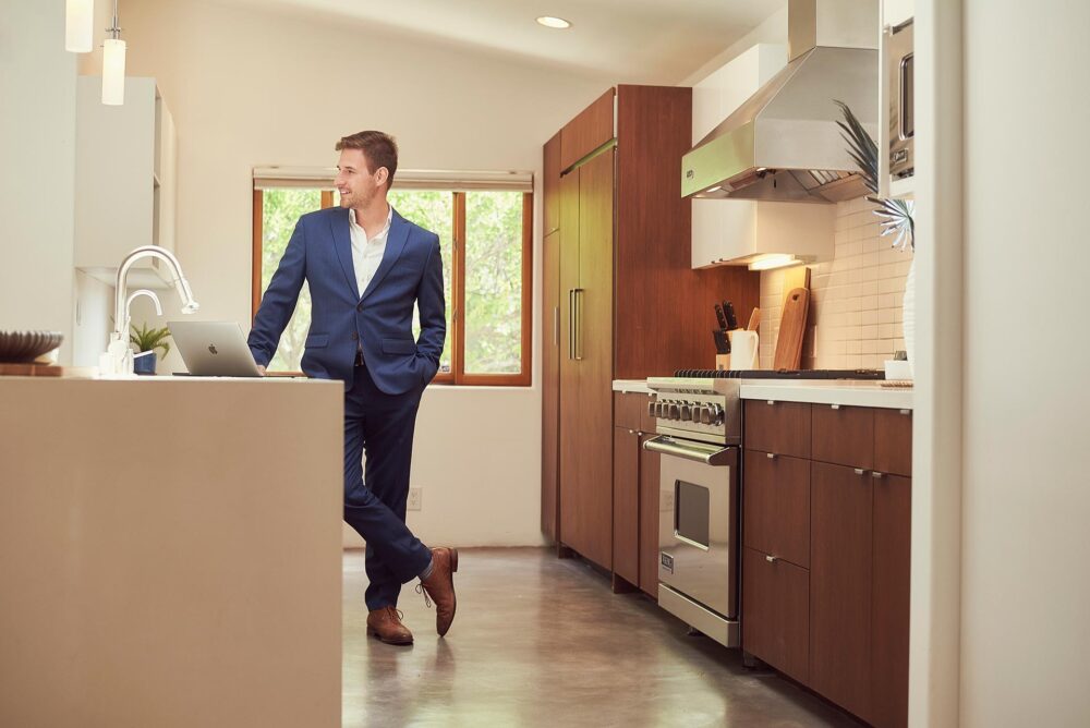 Man in a blue suit standing in a modern kitchen with a laptop, lifestyle dating profile photo with relaxed posture, natural light, and polished wardrobe