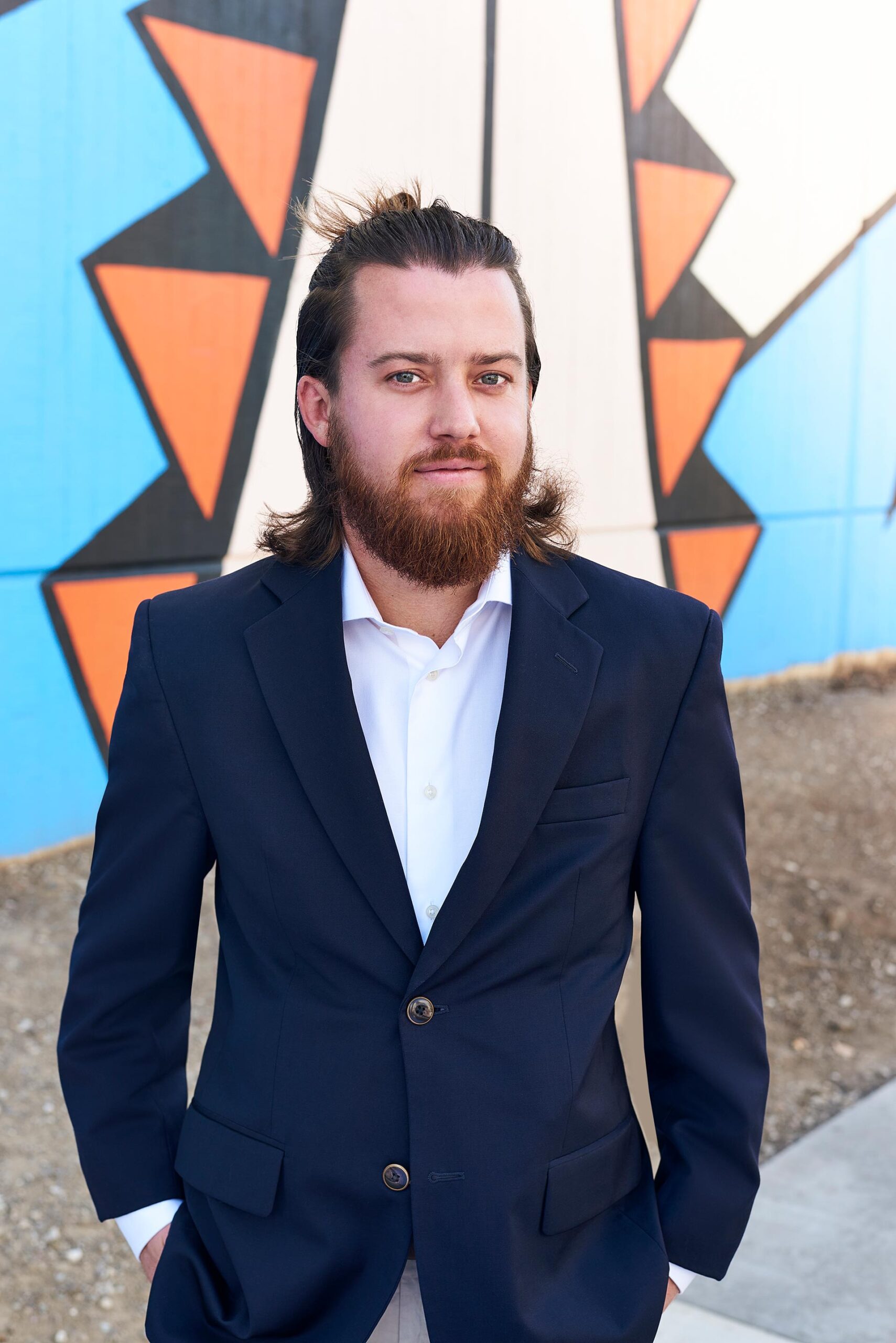 Man in a navy blazer and white shirt standing outdoors in front of a colorful mural, dating profile portrait with direct eye contact and confident relaxed posture