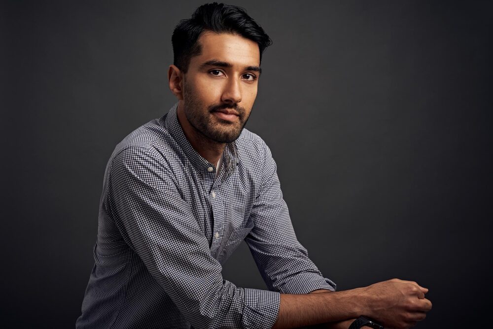 Man seated against a dark studio background, dating profile portrait with checkered button-down shirt, direct eye contact, relaxed posture, and clean lighting