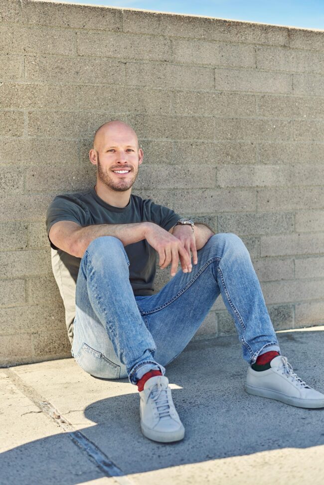 Man seated outdoors against a concrete wall, casual dating profile portrait with green T-shirt, jeans, white sneakers, natural smile, and relaxed body language