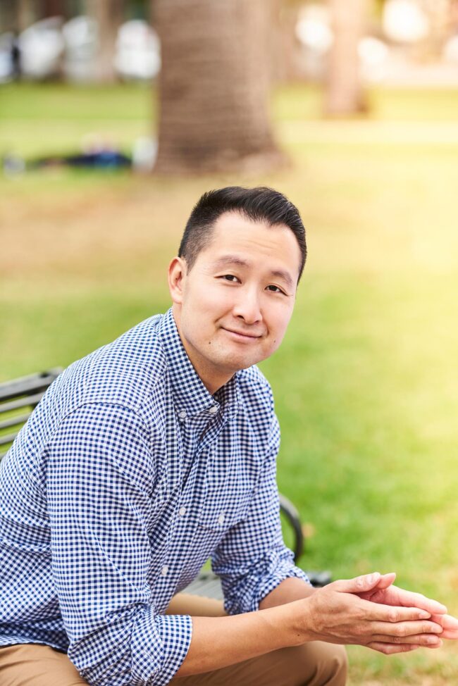 Man seated on a park bench outdoors, dating profile portrait with blue checkered shirt, natural smile, soft greenery, and relaxed body language