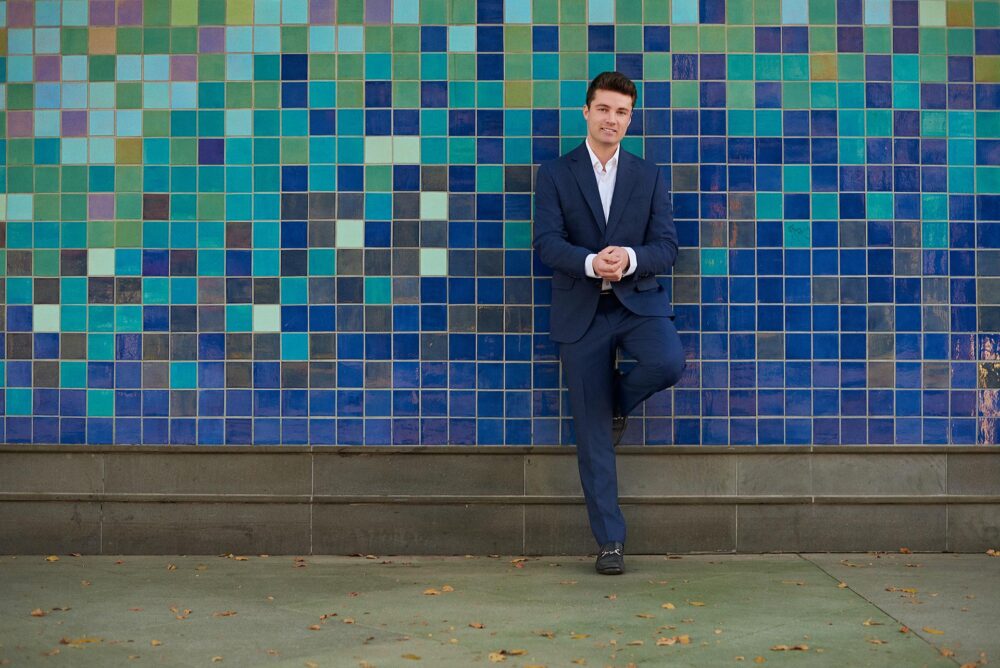 Urban dating profile photo for men in San Francisco, Polished outdoor portrait of a man in a navy suit against a blue tile wall, Confident dating app photo for Hinge Bumble and Tinder, Online dating photographer San Francisco image with clean style and approachable posing.