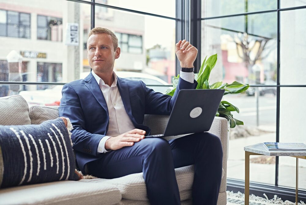 Professional lifestyle dating profile photo for men in San Francisco, Man in a blue suit seated with a laptop in a bright indoor setting, Polished online dating photo for men who want confident approachable profile pictures, Dating app photographer San Francisco image for Hinge Bumble and Tinder profiles.