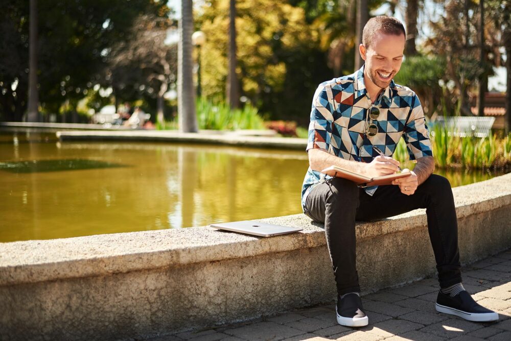 Creative outdoor dating profile photo for men in San Francisco, Man smiling while writing in a notebook near the water, Natural dating app photo for men that shows personality and lifestyle, San Francisco dating photographer image for Hinge Bumble and Tinder profiles.