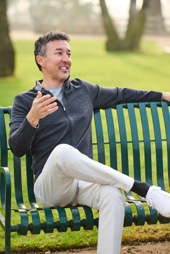 Man seated on a park bench outdoors, casual dating profile portrait with gray zip-up sweater, light pants, natural expression, and relaxed conversational body language