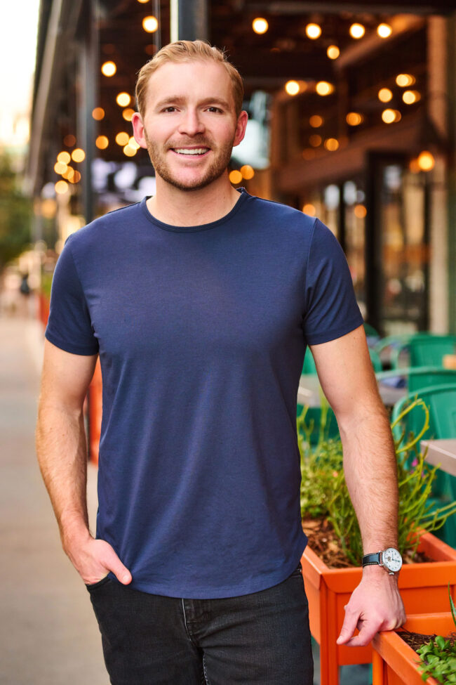 Man standing on a city sidewalk in a navy T-shirt, casual dating profile portrait with natural smile, relaxed posture, warm lights, and outdoor cafe background