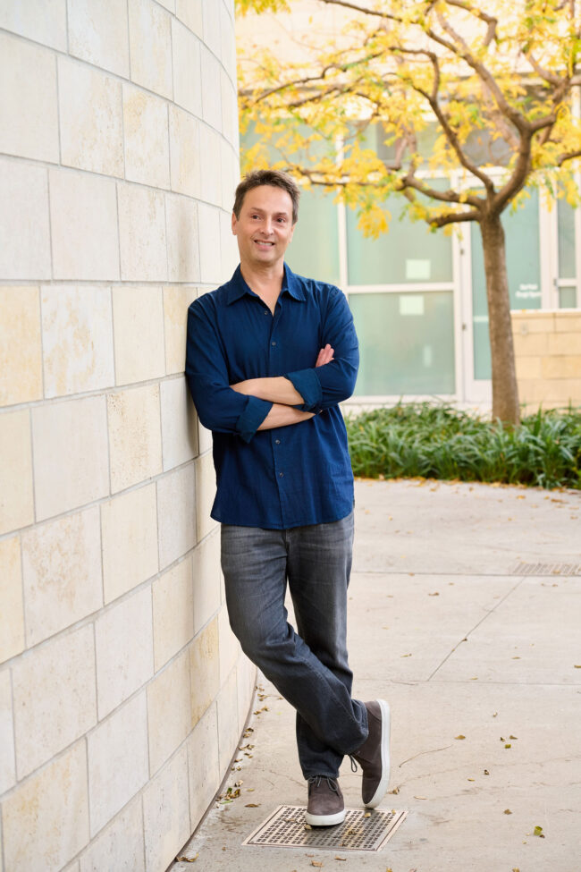 Man leaning against a stone wall in an urban courtyard, full-body dating profile portrait with blue button-down shirt, relaxed crossed-arm pose, greenery, and natural light