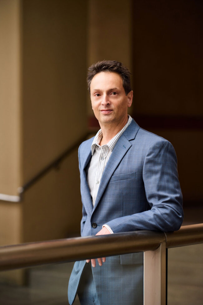 Man in a light blue suit standing indoors by a railing, polished dating profile portrait with direct eye contact, soft interior light, and composed posture