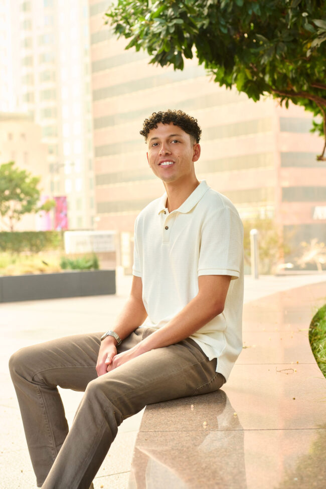 Man seated outdoors in a light polo shirt, casual dating profile portrait with soft city light, relaxed posture, natural smile, and urban background