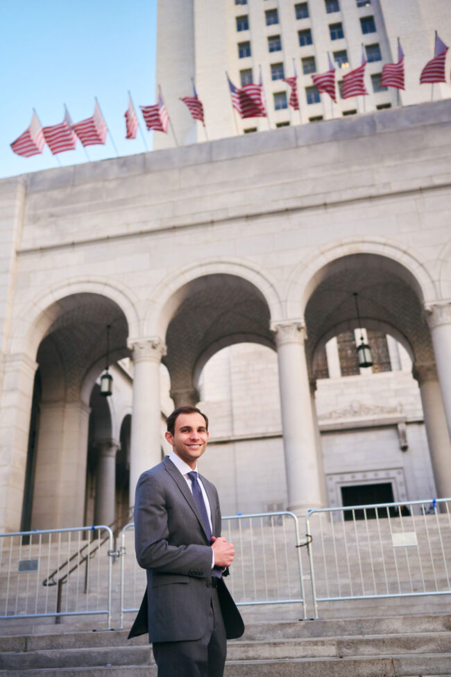 Man in a gray suit standing outside a civic building with columns and flags, polished dating profile portrait with relaxed smile, formal wardrobe, and architectural background