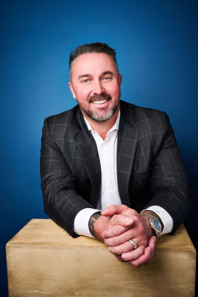 Man seated against a blue studio background, polished dating profile portrait with checkered blazer, white shirt, warm smile, and relaxed hand placement