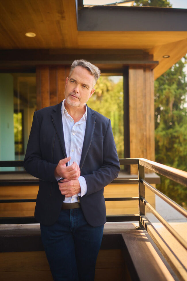Man standing on a balcony outside a modern home, polished dating profile portrait with navy blazer, warm natural light, relaxed pose, and residential background