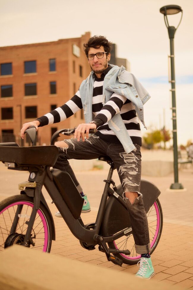 Man wearing a striped sweater, denim jacket, and sneakers while sitting on a bike outdoors.