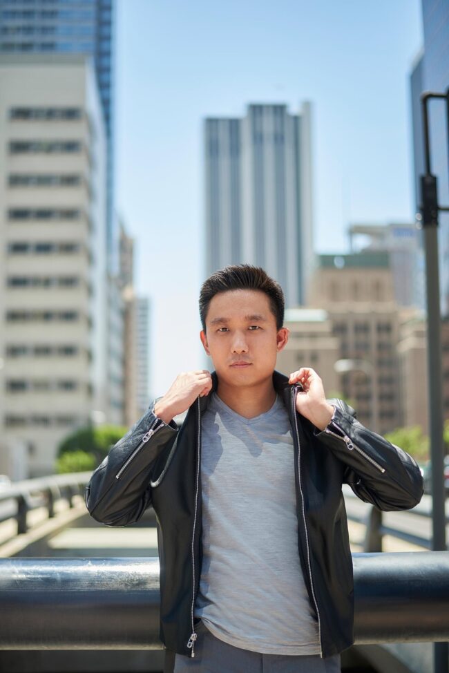 Man wearing a black leather jacket and gray shirt in an urban outdoor dating profile photo.
