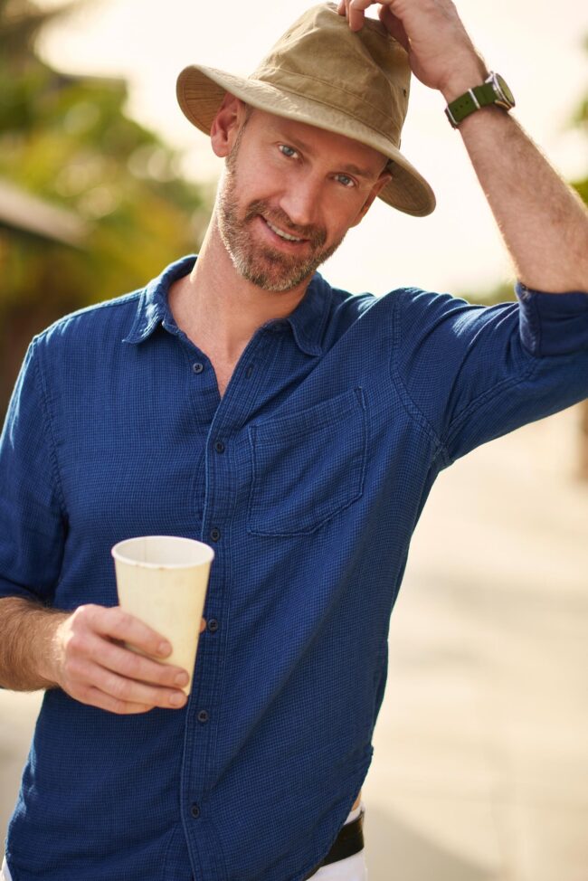 Man wearing a blue button-down shirt and hat while holding coffee in a casual outdoor dating profile photo.
