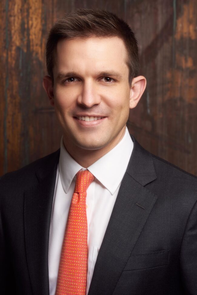 Professional headshot in Los Angeles, man in a dark suit and orange tie photographed against a warm studio background, useful for website bio, LinkedIn profile, resume, company profile, and professional use