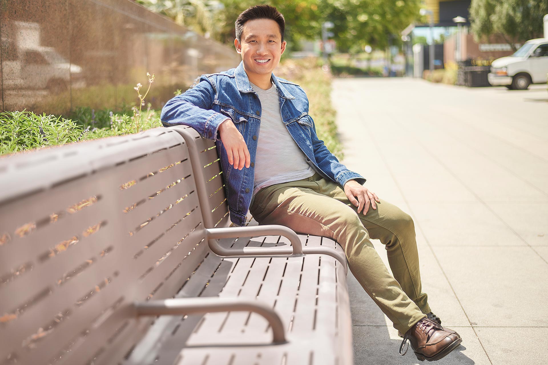Relaxed Profile Photo for First Date Duration Man in a denim jacket sitting on an outdoor bench along a sunny sidewalk.