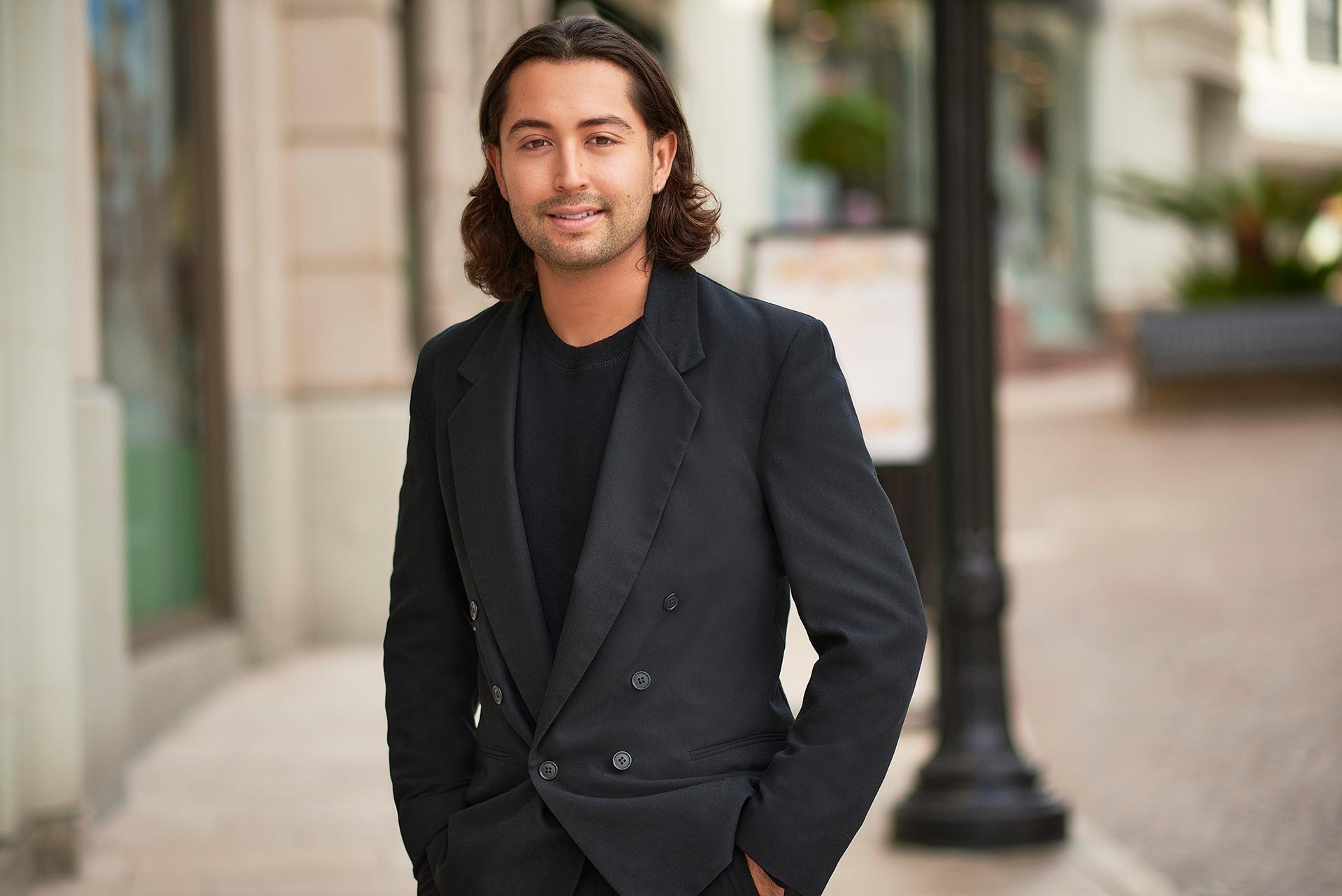 Natural Profile Portrait for First Date Length Man wearing a black blazer and black shirt standing outdoors on a city sidewalk with a softly blurred background.