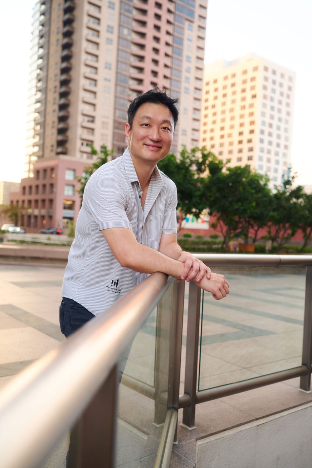 Man leaning on an outdoor railing with city buildings in the background, dating profile portrait with relaxed posture, soft evening light, striped shirt, and natural smile