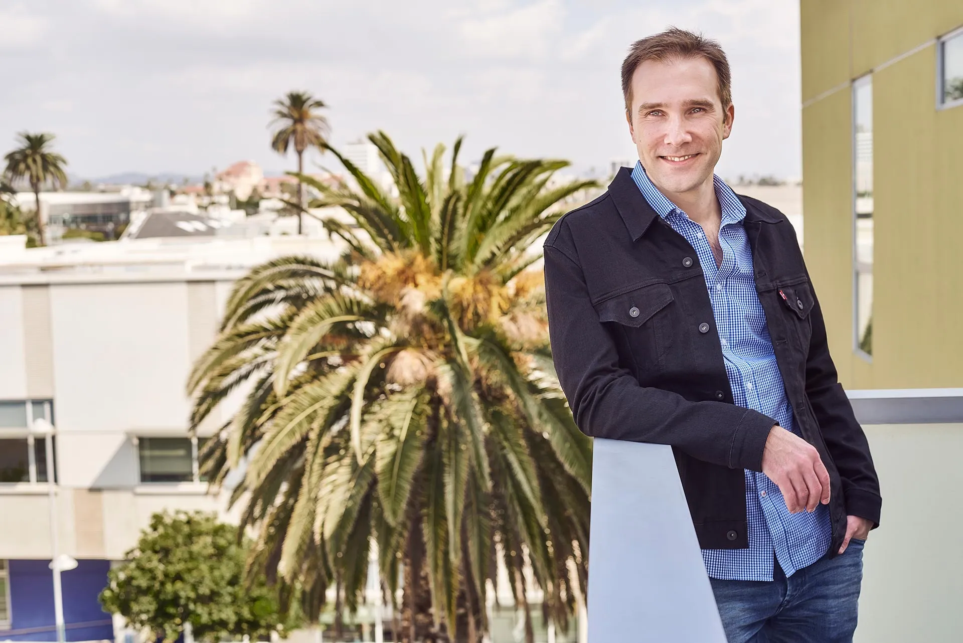 Casual Profile Photo for How to Date in LA Man leaning on a balcony railing, wearing a dark jacket and blue shirt with palm trees and city buildings in the background.