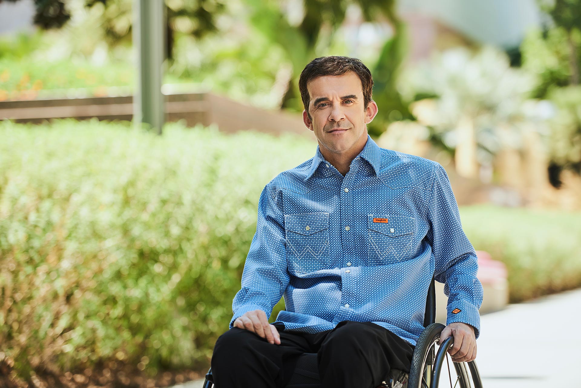 Outdoor Profile Photo for the Los Angeles Dating Scene Man seated in a wheelchair outdoors, wearing a blue button-down shirt with greenery and soft sunlight in the background.