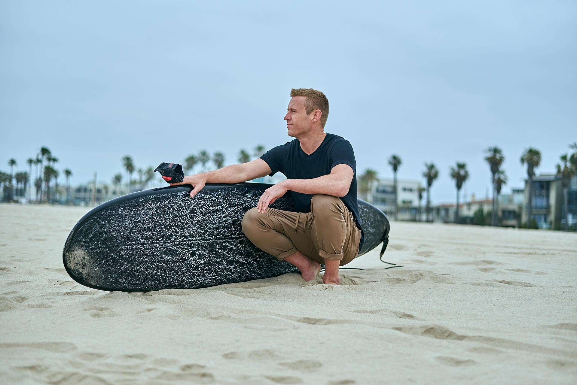 Beach Lifestyle Photo for Single Men in Los Angeles Man kneeling on the sand beside a black surfboard with palm trees and beach homes in the background.